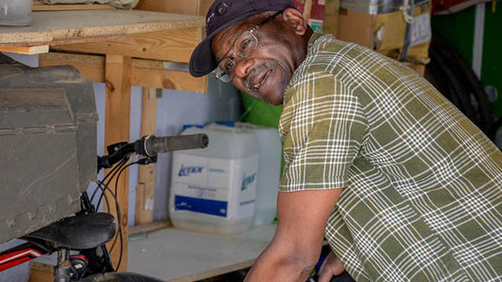 getting-bike-out An older man wearing a baseball hat and glasses. He's in a workshop working on a bicycle.