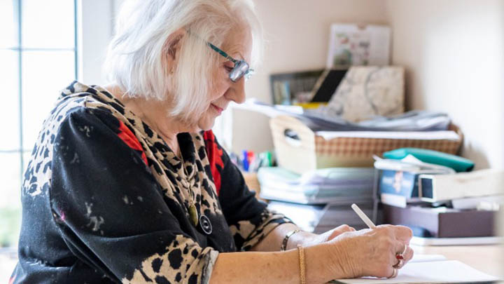 homecare-banner A woman with white hair and glasses. writing with a pen onto paper.