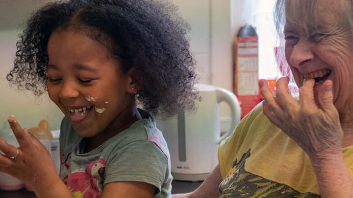 Older woman and grandchildren A child with black curly hair and an older woman with grey hair laughing. They're in a kitchen eating cake batter from their fingers.