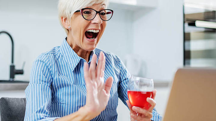A joyful older woman with white hair, wearing glasses and a blue striped shirt, waves at a laptop screen while holding a glass of red drink, celebrating online.
